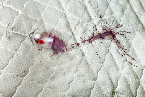 Spilled wine glass on the bed. Accidentally dropped wineglass on white bedsheet. Unlucky, unfortunate situation. Wet stain.