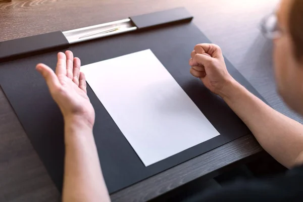 Obchodník sedí u stolu. Na stole je prázdný list papíru. Rozzlobené gesto rukama. Koncept, fotografie. — Stock fotografie