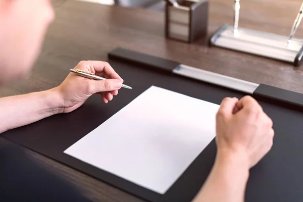Obchodník sedí u stolu a drží pero v levé ruce. Na stole je prázdný list papíru. Rozzlobené gesto rukama. Koncept, fotografie. — Stock fotografie