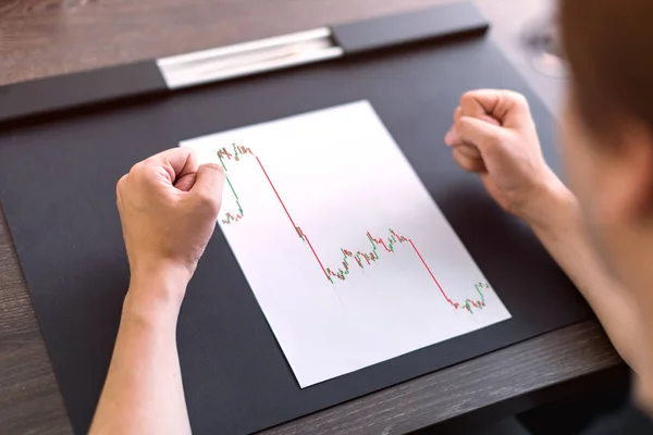 Man sitting at table. There are sheet of paper with a trading chart on the table. Angry gesture with hands. Concept photo.