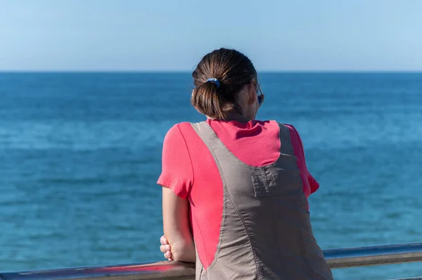 Mujer sueña con viajar y mira al mar. Vista trasera trasera, espacio para copiar. Esperando a un amante o esposo. Clima soleado y mar azul limpio . —  Fotos de Stock