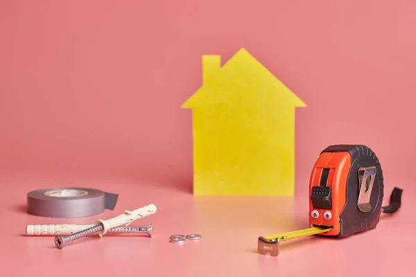 House renovation funny concept. Metal tape measure and other repair items. Home repair and redecorated concept. Yellow house shaped figure on pink background. — Stock Fotó