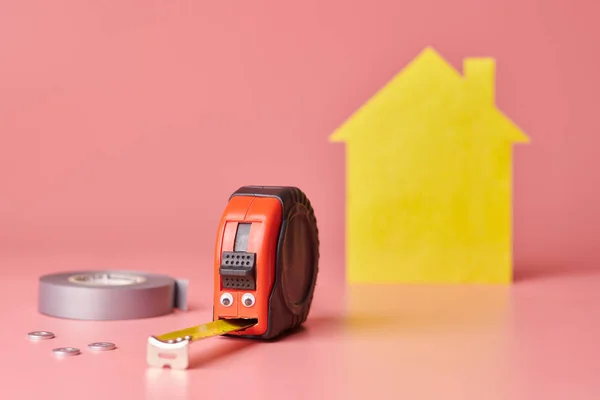 House renovation funny concept. Metal tape measure and other repair items. Home repair and redecorated concept. Yellow house shaped figure on pink background. — Stok fotoğraf
