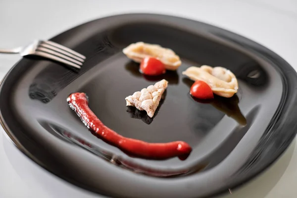 Funny food face, dumpling and ketchup. Food art on black plate. Smiling face on plate. Motivation for losing weight and healthy eating.