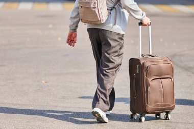 Tekerlekli bavullu yaşlı adam. Havaalanından aşağı doğru yürüyen valizli bir erkek. Seyahat sonrası eve dönüş. Jet lag etkisi. Arka plan