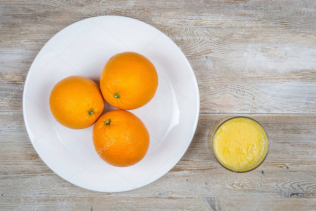 three oranges on a plate next glass with juice on a light wooden background. top view