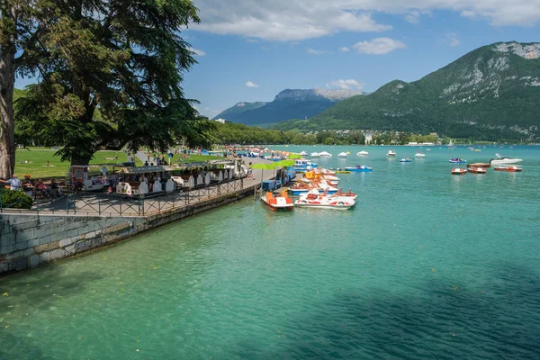 Uma vista ensolarada sobre o Lago Annecy, França, com pedalinhos no cais e turistas no parque — Fotografia de Stock