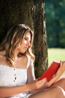 Güzel genç kadın parkta bir ağaç altında bir kitap okuma. 