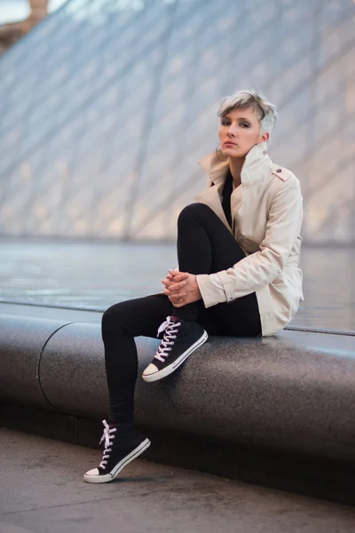 Young woman portrait sit in front of Louvre Pyramid in Paris, France.