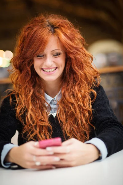 Mujer pelirroja sonriente usando teléfono móvil en la biblioteca pública . —  Fotos de Stock