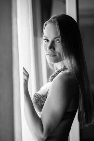 Sensual young woman portrait wearing lingerie at the window in hotel room. Black and white image.