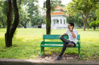 Genç yakışıklı adam telefonda konuşuyor ve parkta bankta otururken gülümsüyor.