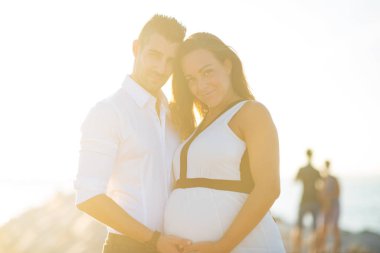 Couple intimate portrait together on the beach in the summer at sunset with natural flare. Man hugging his pregnant woman.