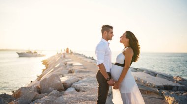 Couple intimate face to face portrait together on the beach in the summer at sunset with natural flare. Man holding hands of his pregnant woman.