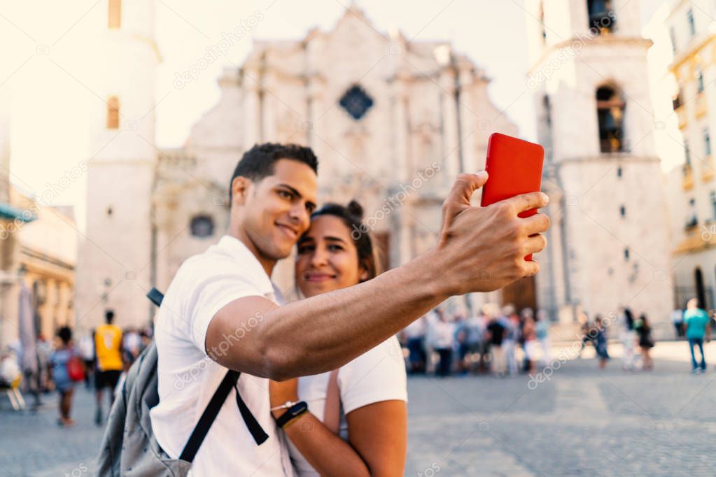 Young smiling couple taking selfie with smartphone in Havana, Cuba. Cathedral in the background.