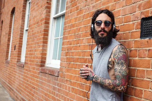 Young tattooed man portrait listening to music against brick wall in Shoreditch borough. London, UK. Hipster style.