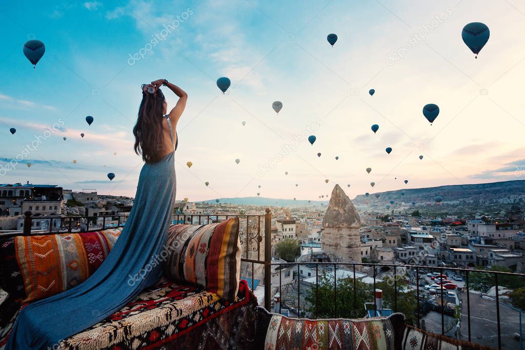 Young beautiful woman wearing elegant long dress in front of Cappadocia landscape at sunshine with balloons in the air. Turkey.
