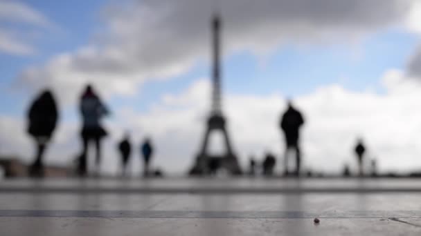 OUT OF FOCUS: Lidé kráčející před Eiffelovou věží proti modrému zamračenému nebi. Paříž, Francie.