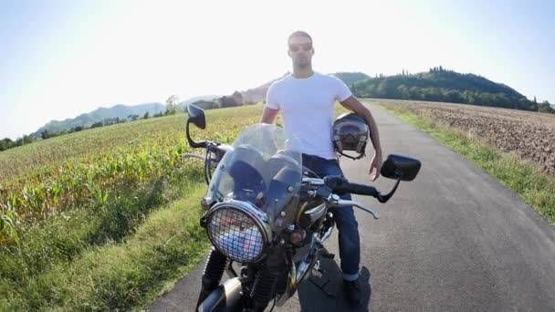 晴れた日に牧草地の中でオートバイにポーズをとる安全ヘルメットと白いTシャツを着たハンサムなスポーティな男 — ストック動画