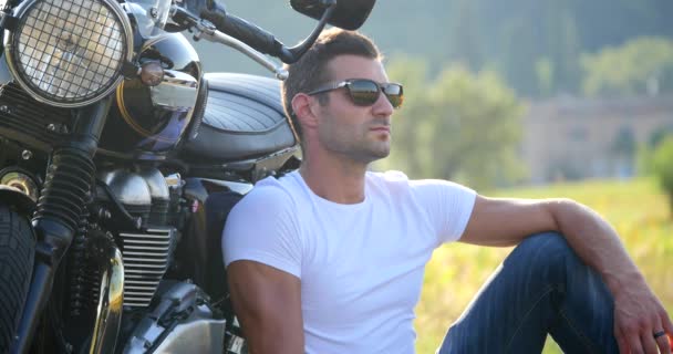 晴れた日に牧草地の中でオートバイの近くでポーズをとる白いTシャツを着たハンサムなスポーティな男 — ストック動画