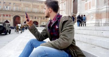 sokakta merdivenlerde oturan ve akıllı telefon kullanan genç adam