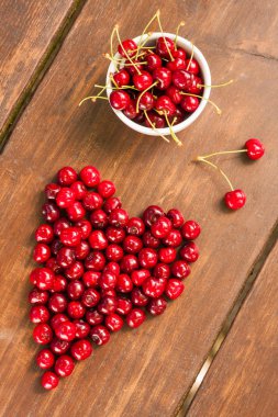 Parlak kırmızı olgun taze erken tatlı kiraz aldım yerleştirilen düzgünce koyu ahşap bir masa üzerinde. Bir kalp - aşk sembolü şekli oluşturan tek kiraz saplarını bir küçük beyaz seramik kase ile meyve.
