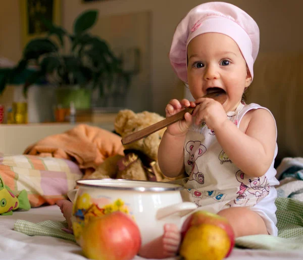 鍋料理とバック グラウンドでいくつかのおもちゃを持ついくつかのリンゴの前に木のスプーンが保持している彼女の頭の上のシェフのキャップとコックとして服を着た女の赤ちゃん — ストック写真