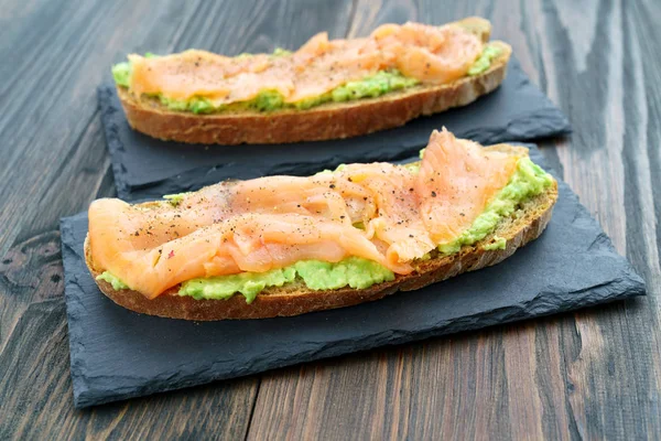 Rye toasts with smoked salmon and mashed avocado served on slate plates. Dark wooden background, high resolution