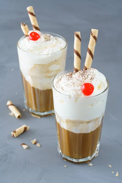 Ice coffee topped with vanilla ice cream, whipped cream, wafer straws and cocktail cherry. Gray table, high resolution.