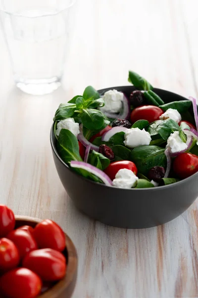 Winter salad with lamb\'s lettuce, spinach, cherry tomatoes, red onion, dried cranberries and goat cheese. Grey ceramic bowl, white wooden table, high resolution
