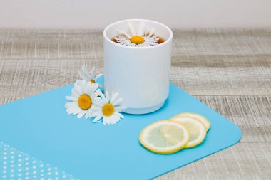 papatya çiçekleri, dilimlenmiş limon ve gri ahşap bir masa üzerinde polka nokta lı bez ile mavi bir kesme tahtası üzerinde papatya çayı ile beyaz fincan