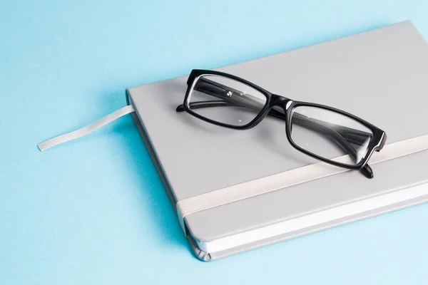 black-rimmed glasses lie on a black leather notebook, blue background, copy space