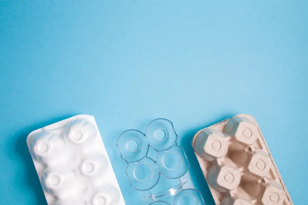 foam, plastic and cardboard egg packaging on a blue background, top view, copy space, various boxes and a reusable plastic egg stand for storage in the refrigerator