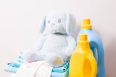 Açık sarı ve mavi renkli plastik şişelerde jel ve kumaş yumuşatıcı, renkli çocuk kıyafetleri ve bir oyuncak yığını, parlak bir arkaplanda kurutmak için mandallar.