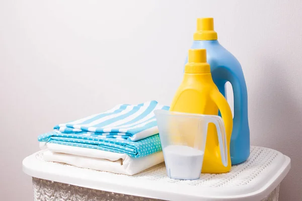 a stack of colored laundry, detergent in a measuring cup and washing gels in yellow and blue bottles on a white plastic basket for dirty laundry, copy space