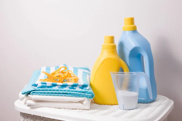 a stack of colored laundry, clothes pegs, laundry detergent in a measuring cup and washing gels in yellow and blue bottles on a white plastic basket for dirty laundry, copy space