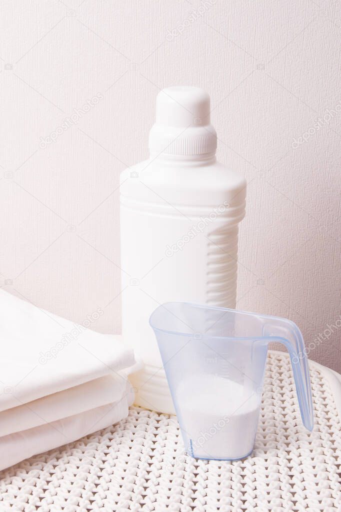 white plastic bottle with bleach and laundry detergent in a measuring cup on a white plastic basket for dirty laundry, light background copy space, wash white linen concept
