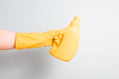 Sarı lastik eldivenli kadın eli açık mavi arka planda etiketsiz sarı bir çamaşır jeli tutar.