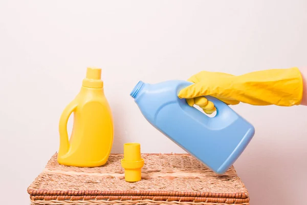 a female hand pours a washing gel for washing children\'s clothes in a measuring cap, a washing gel and a fabric softener on a wicker basket, light background, copy space