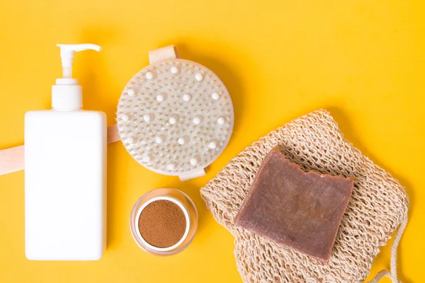 a jar of ground coffee, a brush for dry anti-cellulite massage, a knitted washcloth, homemade cocoa soap,, a white bottle with a dispenser without a label on a yellow background, body care at home