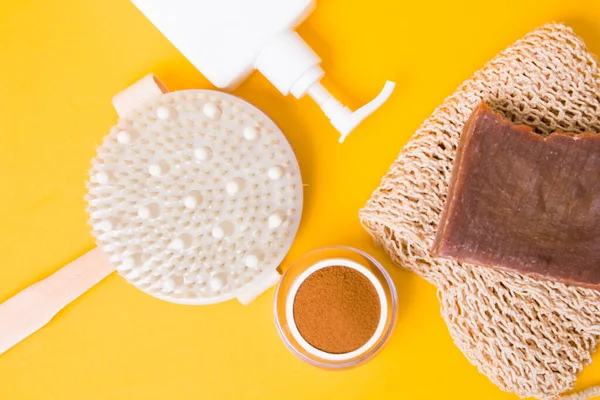 a jar of ground coffee, a brush for dry anti-cellulite massage, a knitted washcloth, homemade cocoa soap,, a white bottle with a dispenser without a label on a yellow background, body care at home