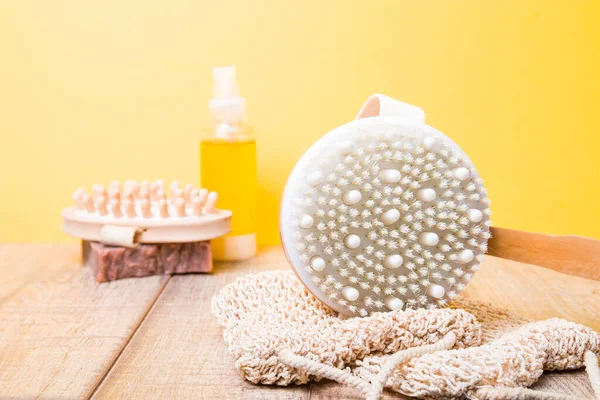 a brush for dry anti-cellulite massage on a wooden table, a knitted washcloth, homemade cocoa soap, a wooden massager and body care oil on a yellow background