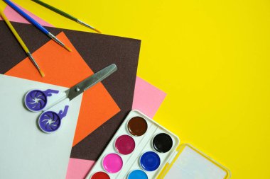 Okul konseptine, boya fırçasına, sulu boya boyaya, makasa ve sarı arka planda kadife renkli kağıda, kopyalama alanına, üst manzaraya