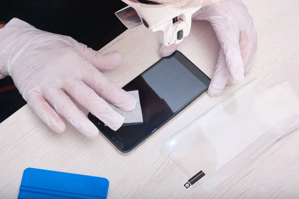 a man in rubber gloves wipes the smartphone glass with a damp cloth before sticking on the protective glass, checking for dust through a magnifying glass