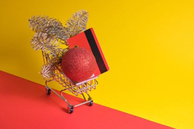 Kırmızı kredi kartı, köknar dalı ve küçük alışveriş arabasında noel ağacı topu kırmızı ve sarı arka planda, yeni yıl alışveriş konsepti