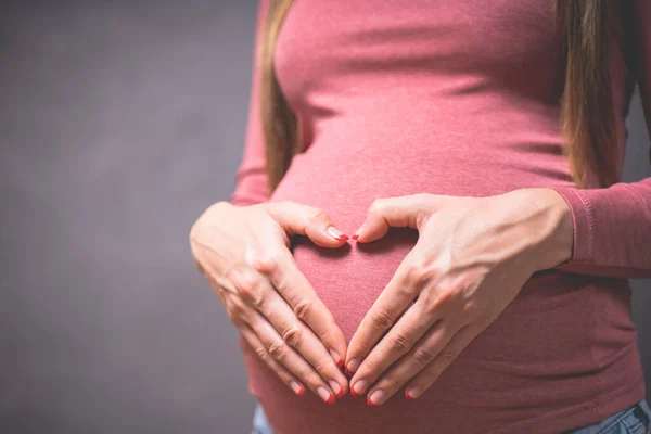 Hamile bir kadın bebeğinin karnında elini kalp şeklinde tutuyor. Parmakları kalp sembolü olan hamile bir karın. Annelik konsepti. Hamileliğin tonlu fotoğrafı. — Stok fotoğraf
