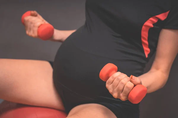 Young pregnant woman exercises with red fitball and dumbell. Working out and fitness, pregnancy concept. Fitness on last months of pregnancy. Active and sportive pregnancy, healthy motherhood concept