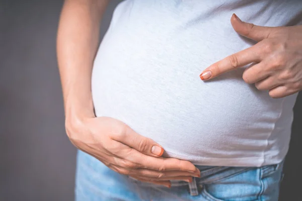 Hamile bir kadın gri bir arka planda el ele tutuşuyor. Hamilelik, annelik, insanlar ve beklenti konsepti. Yakın plan, fotokopi odası, kapalı alan. Hamileliğin tonlu fotoğrafı. — Stok fotoğraf