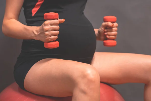 Young pregnant woman exercises with red fitball and dumbell. Working out and fitness, pregnancy concept. Fitness on last months of pregnancy. Active and sportive pregnancy, healthy motherhood concept