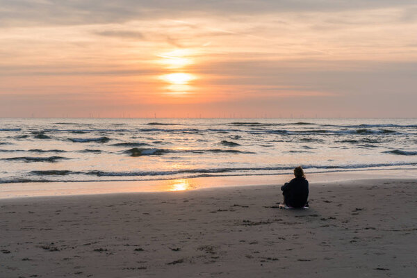 Egmond-aan-Zee, Netherlands, 09-24-2016: Young woman sitting on the beach and looking out over the North Sea and the far offshore energy windmills at sunset. - evening meditation. Egmond aan Zee, the Netherlands